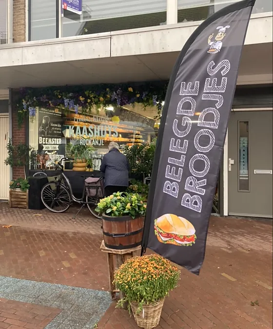 De winkel aan het Dorpsplein met bloemen boven het raam en de vlag
